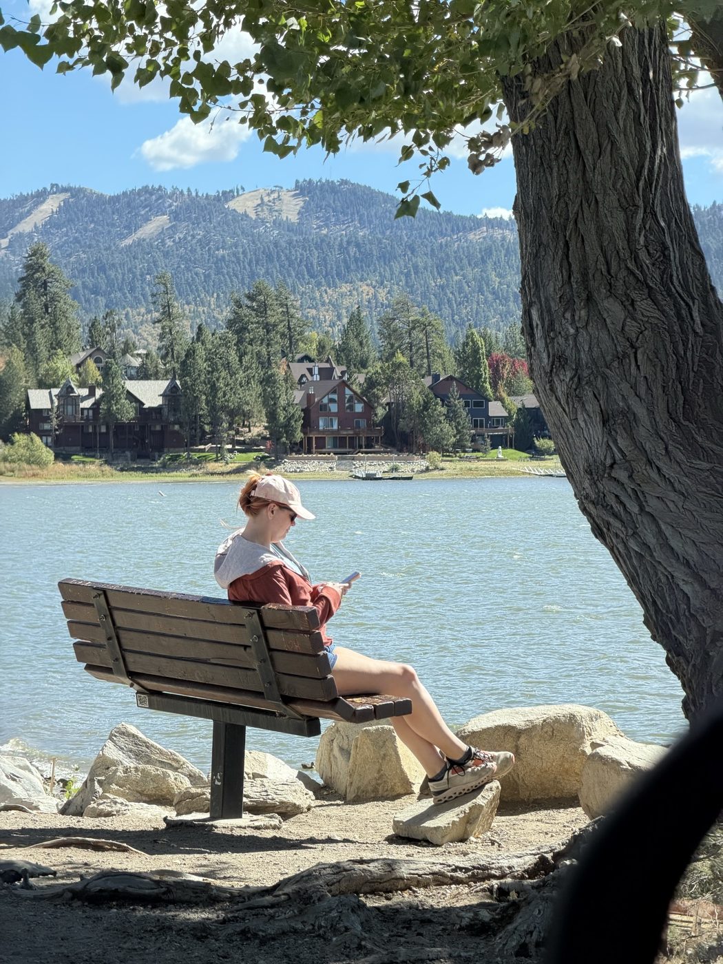 Jenna on a bench by the lake, with big cabins and a forested ski hill across the water