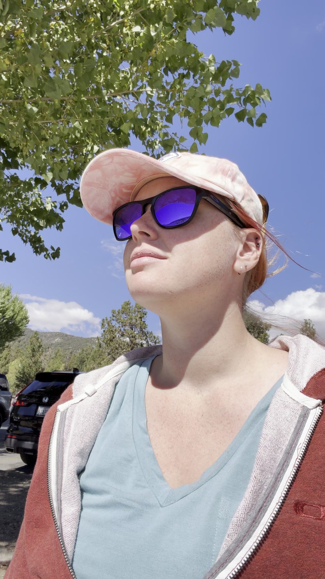 Jenna in a cap, sunglasses and a zip-up hoodie under the trees