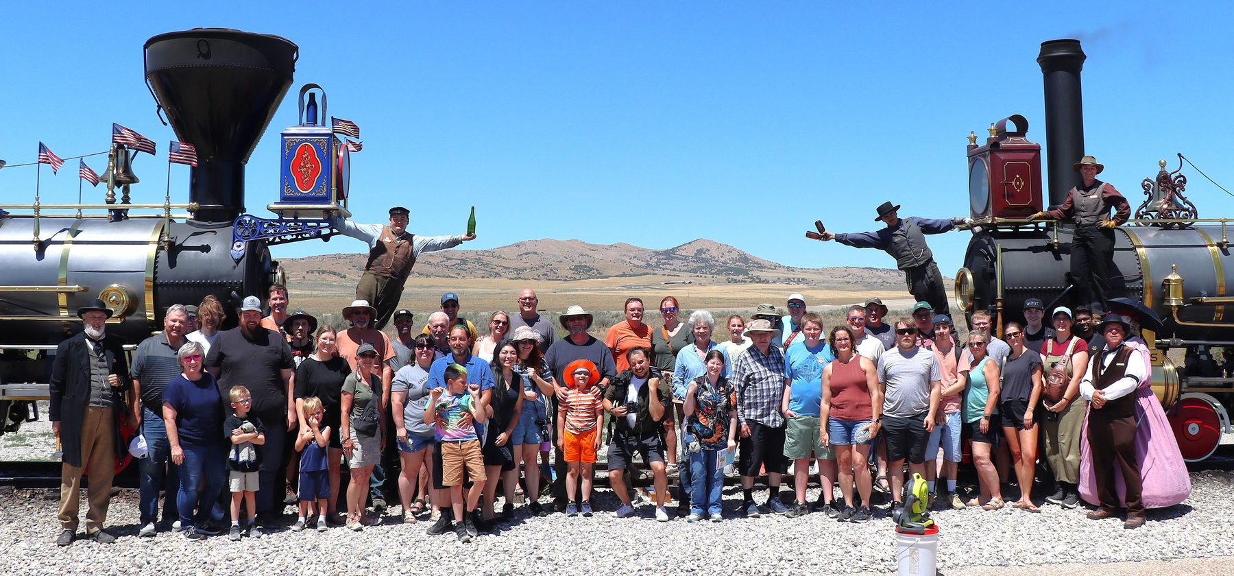 A large group of visitors lined up between two nineteenth century locomotives, with costumed re-enactors standing on the engines, recreating the 1869 photograph