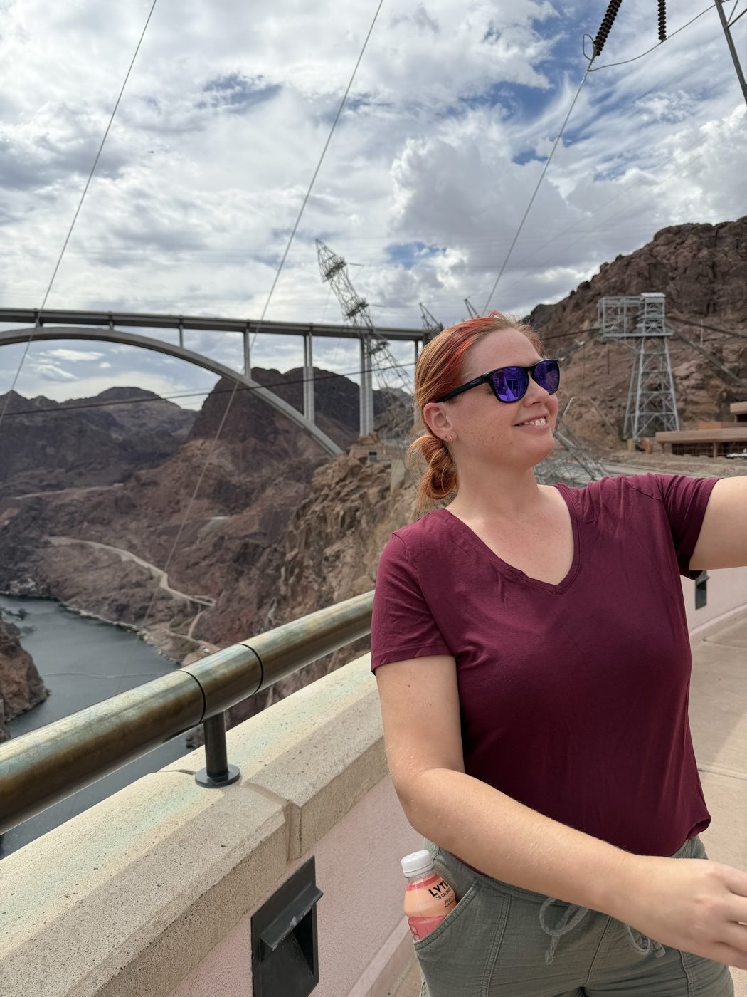 Jenna in sunglasses on top of the dam, with the arch bridge and the canyon behind her
