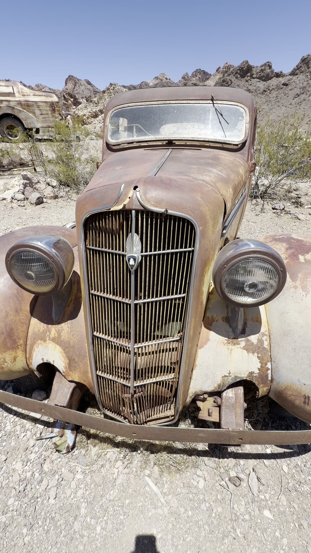 The rusted front of a 1930s car, grille and headlamps intact, sitting on gravel below desert hills