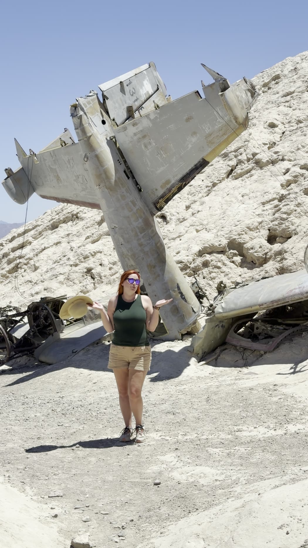Jenna in a green top and shorts posing in front of a crashed plane nose-down in a pale hillside
