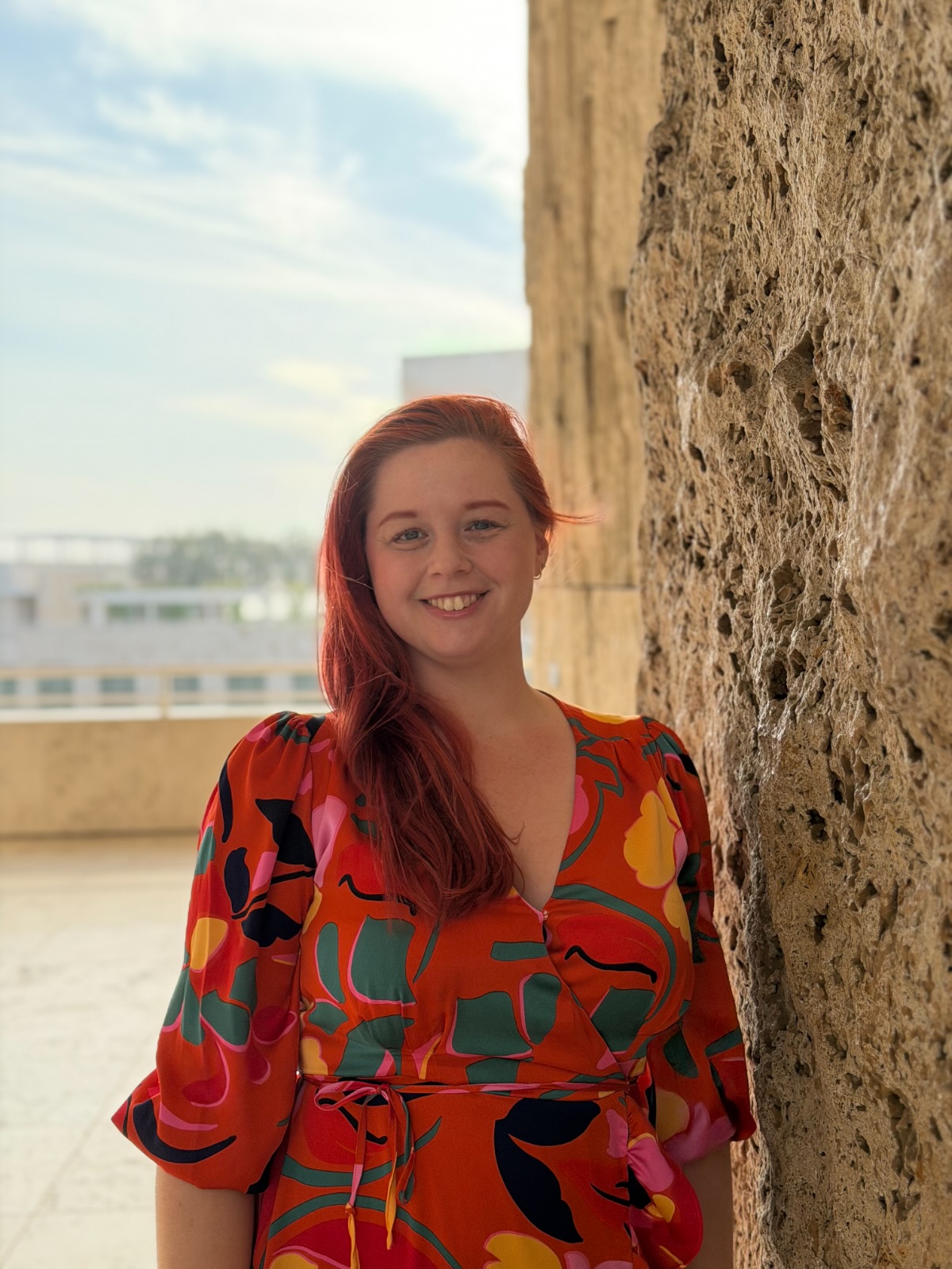 Jenna Harbour, smiling, long red hair, in an orange patterned dress beside a pale stone wall