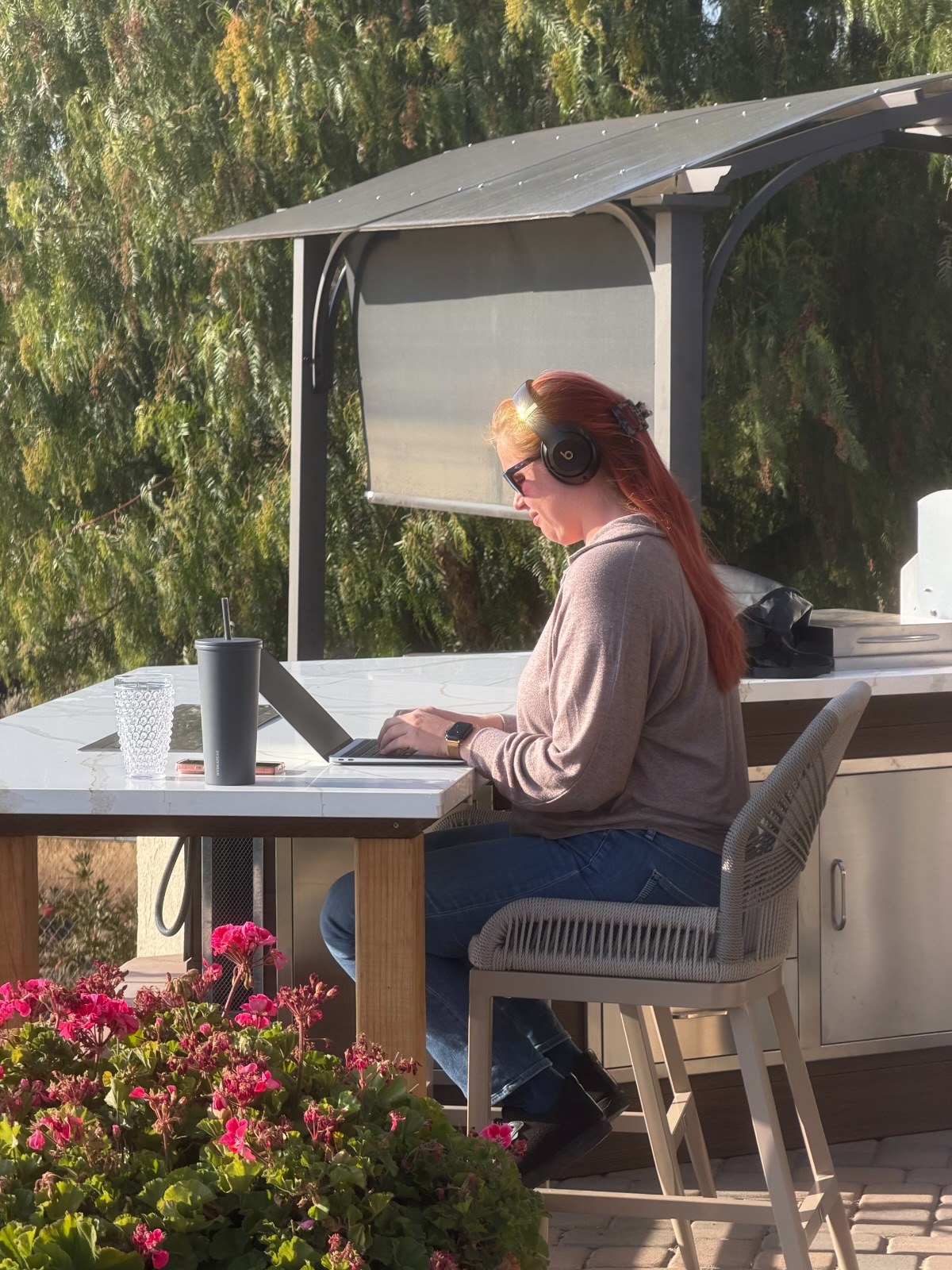Jenna in headphones, typing on a laptop at an outdoor counter, with pink geraniums in the foreground
